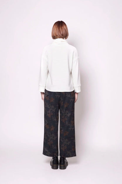 Back view of a woman wearing the Sidney Top by Tangente in Ivory, a slightly cropped, boxy top in ribbed fabric with long cuffed sleeves and a large cowl neck. She is wearing it with floral pants and standing in front of a white wall. 