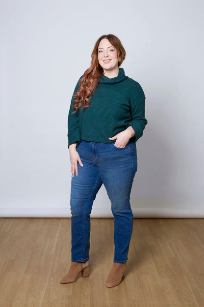 A woman wearing the Sidney Top by Tangente in Pine, a slightly cropped, boxy top in ribbed fabric with long cuffed sleeves and a large cowl neck. She is wearing it with jeans and standing in front of a white wall. 