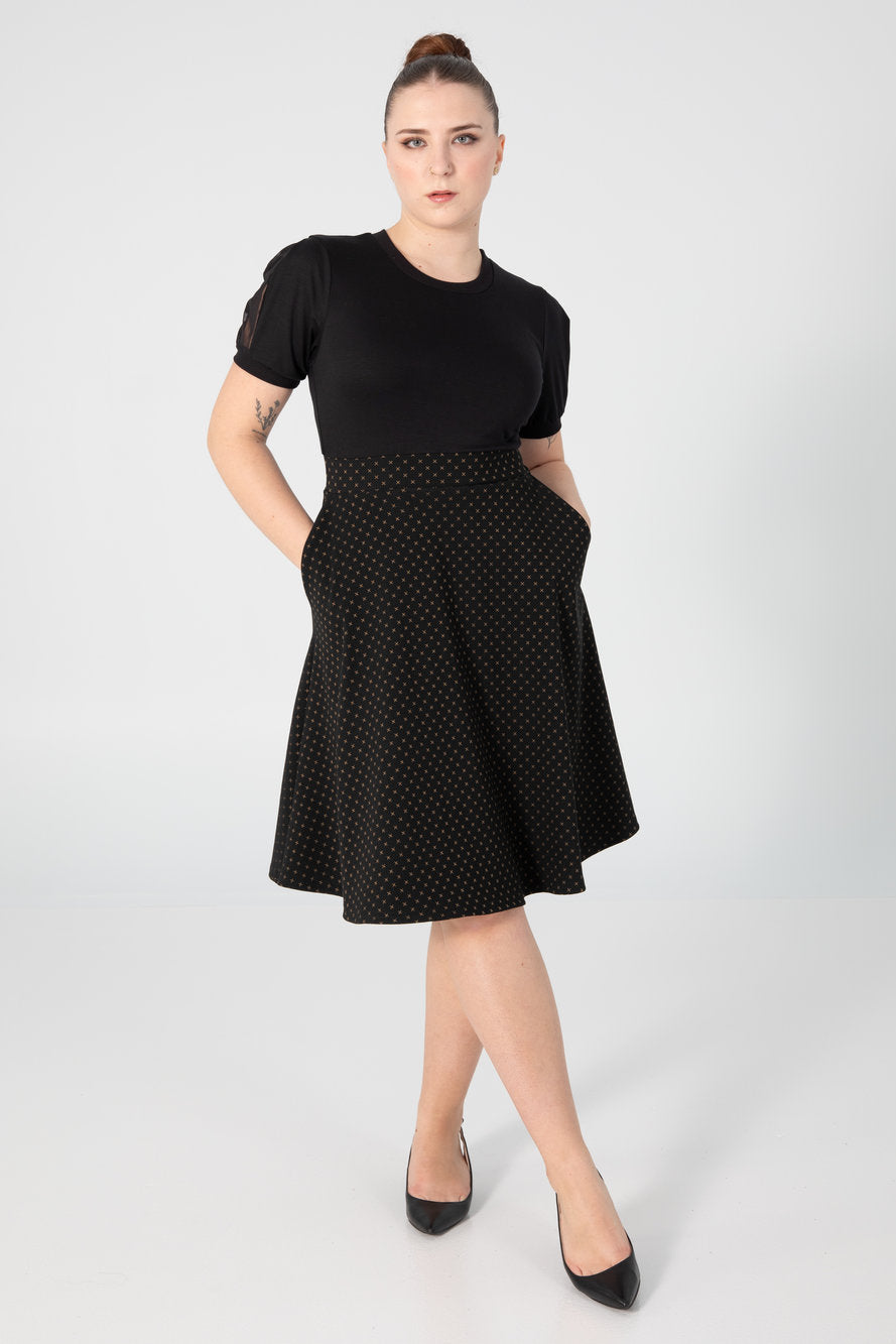 A woman wearing the Darling Dress by Slak with a black bodice with short sleeves with a sheer polka dot inset, a fitted waist, and a flared below-the-knee length skirt in a Golden X on black pattern. 