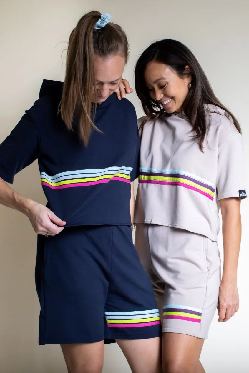 A woman wearing the Striped Bamboo Short by Olive+Splash, a medium length pull-on short in navy with bright stripes across one thigh. She is wearing it with the Bamboo T-Shirt hoodie and standing beside a woman wearing the same outfit in beige. 