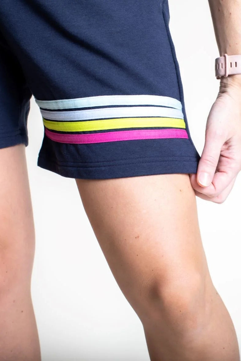 Close up detail of the Striped Bamboo Short by Olive+Splash, a medium length pull-on short in navy with bright stripes across one thigh. She is standing in front of a white background. 
