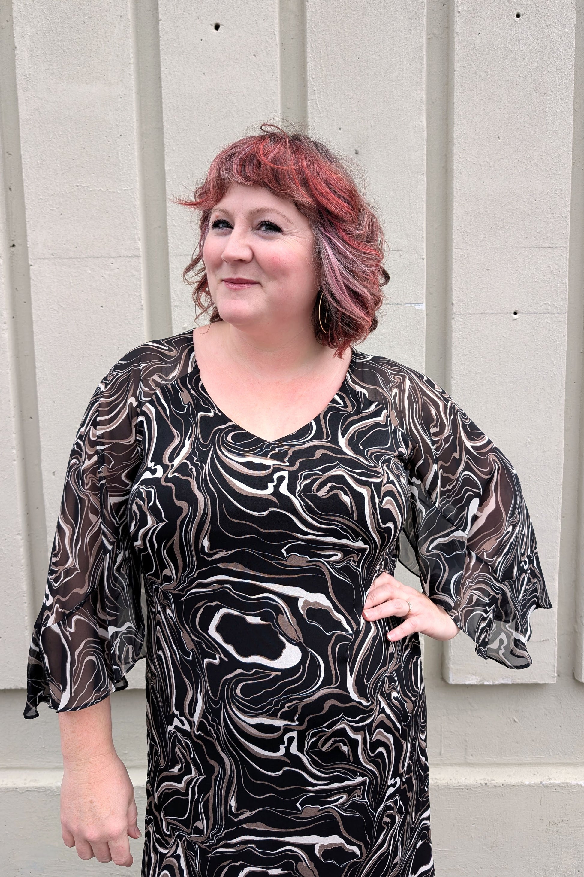 A woman wearing the Nyla Dress by Compli K in Black/Beige Swirl, with a V-neck, fly-away chiffon sleeves with a slit detail, sheath shape, and above the knee hem. She is standing outside in front of a white wall. 