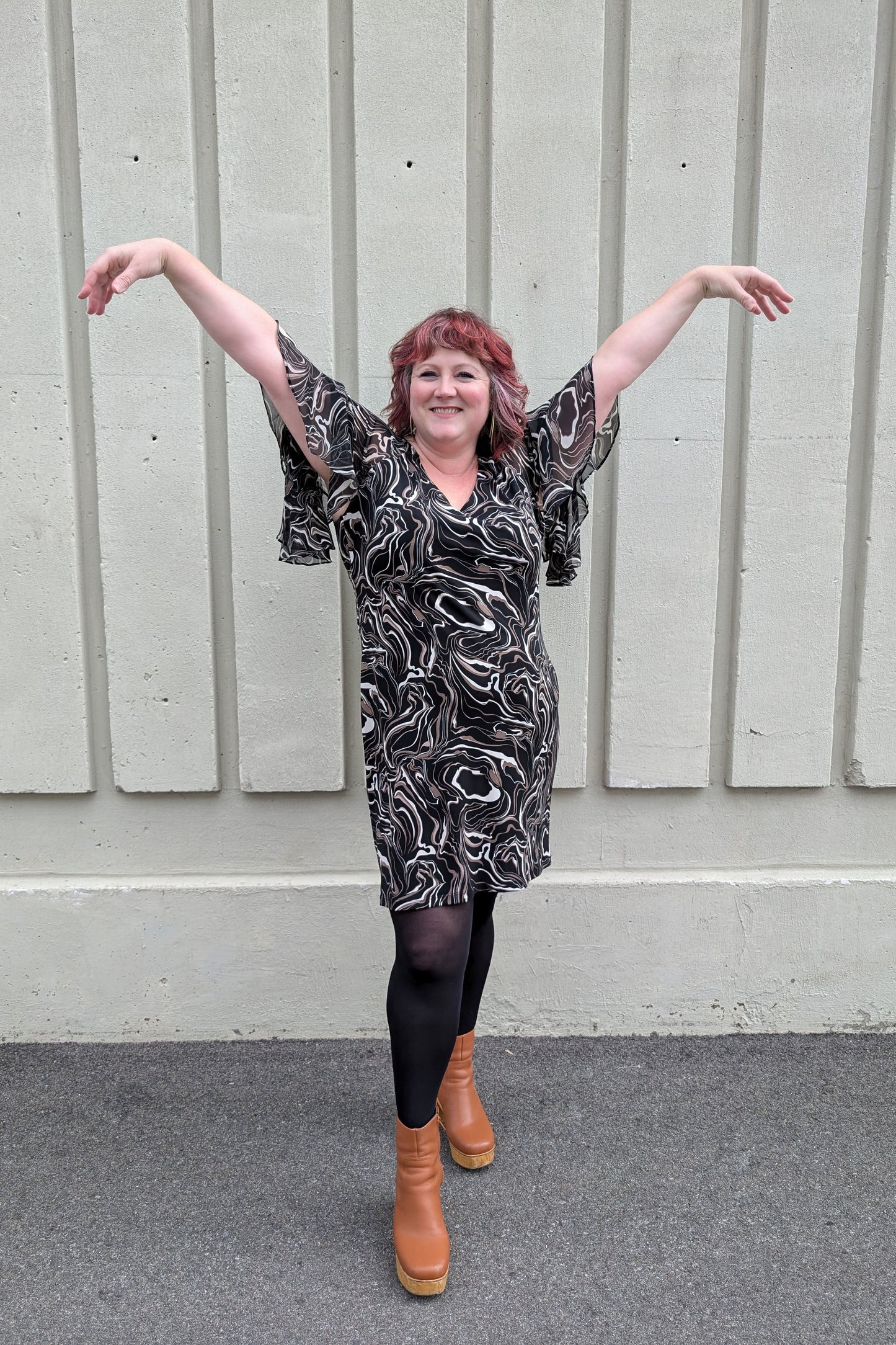 A woman wearing the Nyla Dress by Compli K in Black/Beige Swirl, with a V-neck, fly-away chiffon sleeves with a slit detail, sheath shape, and above the knee hem. She is standing outside in front of a white wall. 