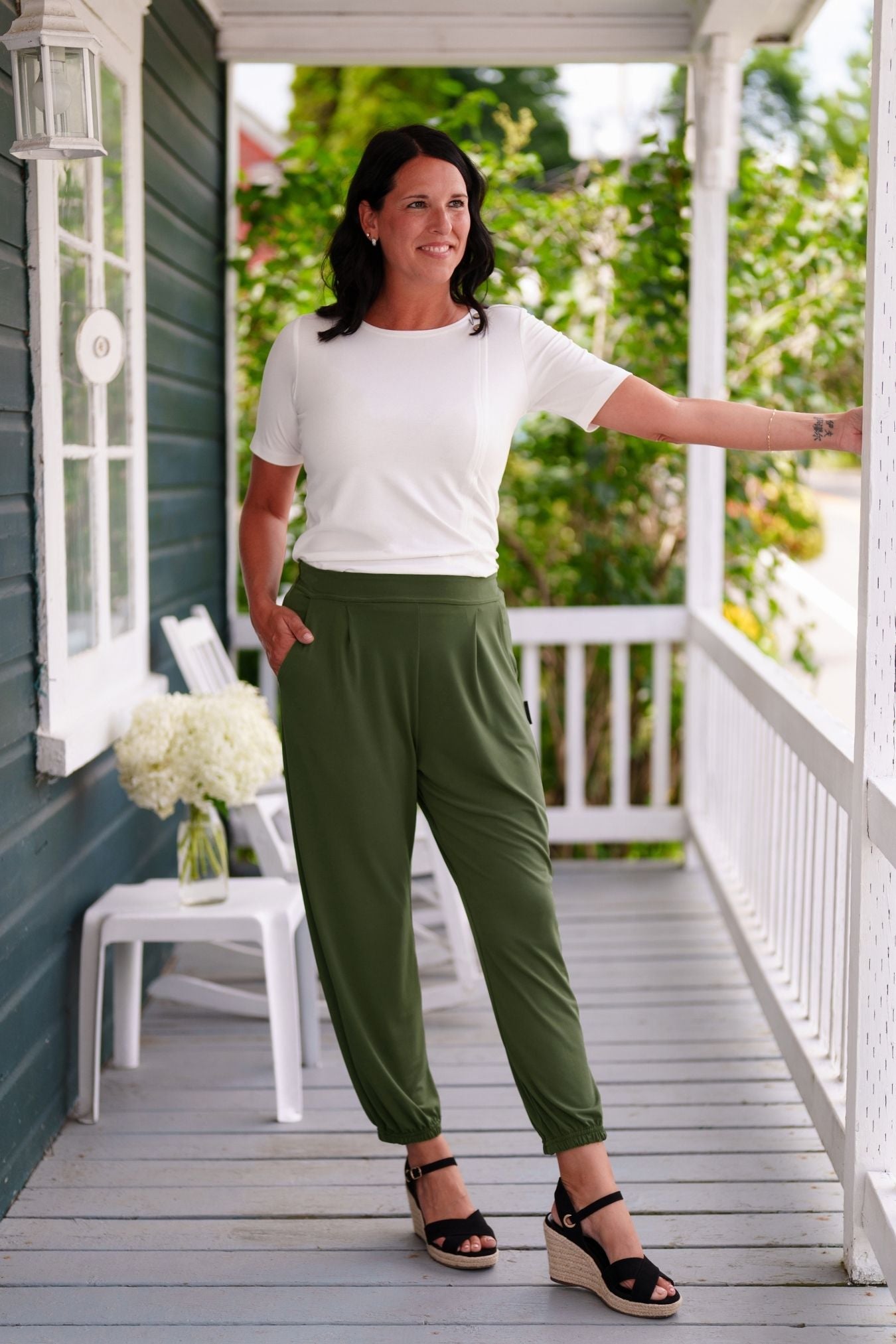 A woman wearing the Mai Tai T-shirt from Rien ne se Perd in Ivory, with coconut buttons at the shoulder, seam accents front and back, and a rounded hi-low hemline. She is wearing it with green pants and standing on a wooden porch. 