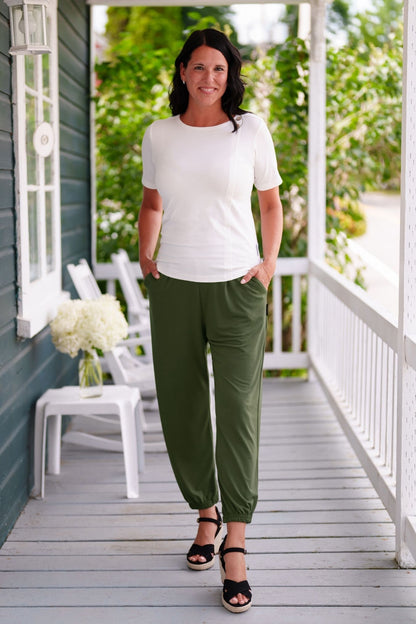 A woman wearing the Mai Tai T-shirt from Rien ne se Perd in Ivory, with coconut buttons at the shoulder, seam accents front and back, and a rounded hi-low hemline. She is wearing it with green pants and standing on a wooden porch. 