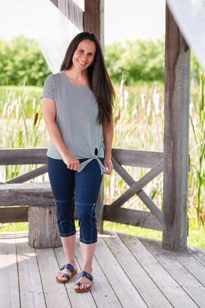 A woman wearing the LIndy Capri pants from Rien ne se Perd, faux denim stretch capris with a wide pull-on waist, contrast seams at the bottom, and decorative coconut buttons. She is wearing them with a striped t-shirt aand standing in a wooden gazebo. 