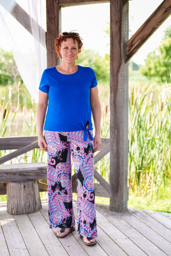 A woman wearing the Modern T-Shirt by Rien ne se Perd in Blue, a bamboo tee with a round neck, short-sleeves, and a tie at the waist. She is wearing them with patterned palazzo pants and is standing outside in a wooden shelter. 