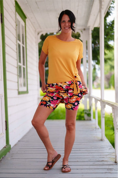 A woman wearing the Cocktail Skort by Rien ne se Perd in Red/Orange floral print, an elastic waist miniskirt with built-in matching bike shorts. She is wearing it with a yellow t-shirt and standing on a porch.