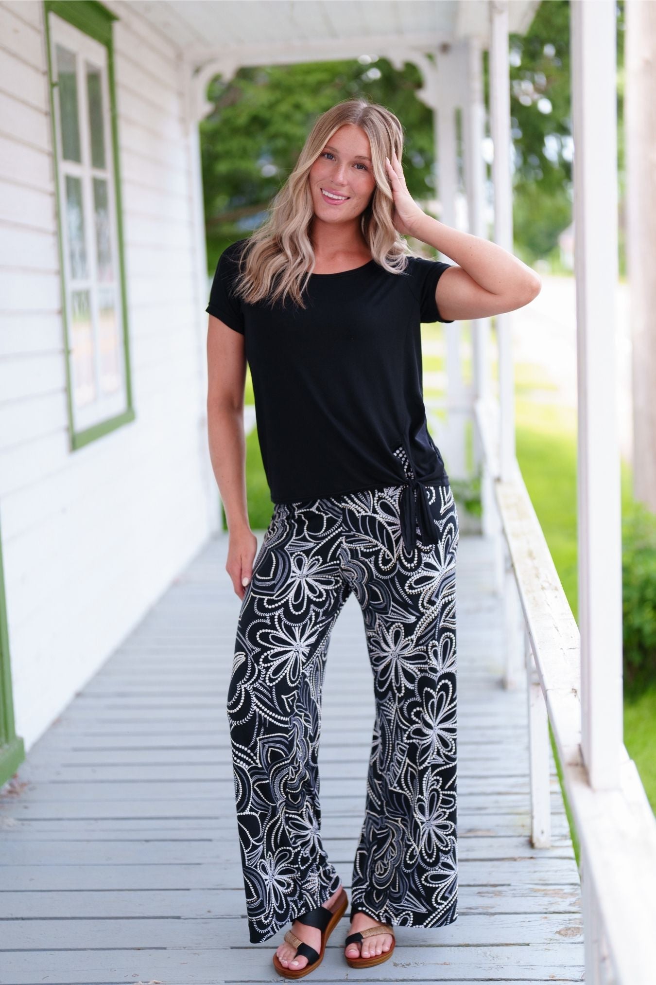 A woman wearing the Coco Palazzo Pants by Rien ne se Perd in Black and White floral, with a wide elastic waistband and wide legs. She is wearing them with a black top and standing on a wooden porch.