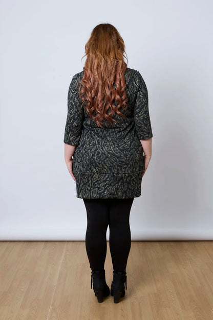 Back view of a woman wearing the Brooke Tunic by Tangente in Green Print, with 3/4 sleeves and a fitted band at the bottom. She is wearing it with black leggings and standing against a white wall. 