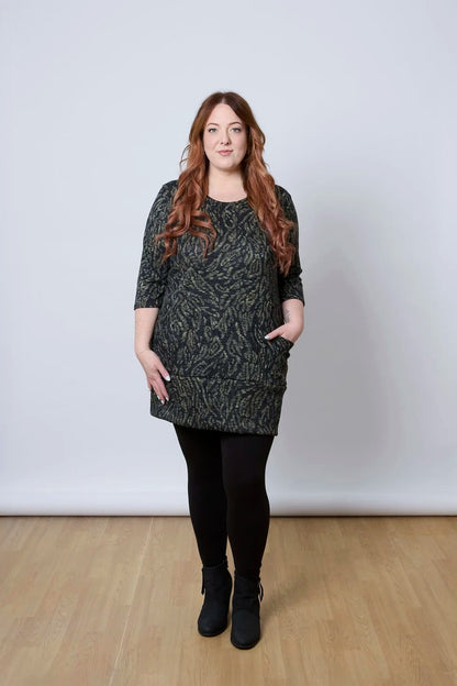 A woman wearing the Brooke Tunic by Tangente in Green Print, with 3/4 sleeves and a fitted band at the bottom. She is wearing it with black leggings and standing against a white wall. 