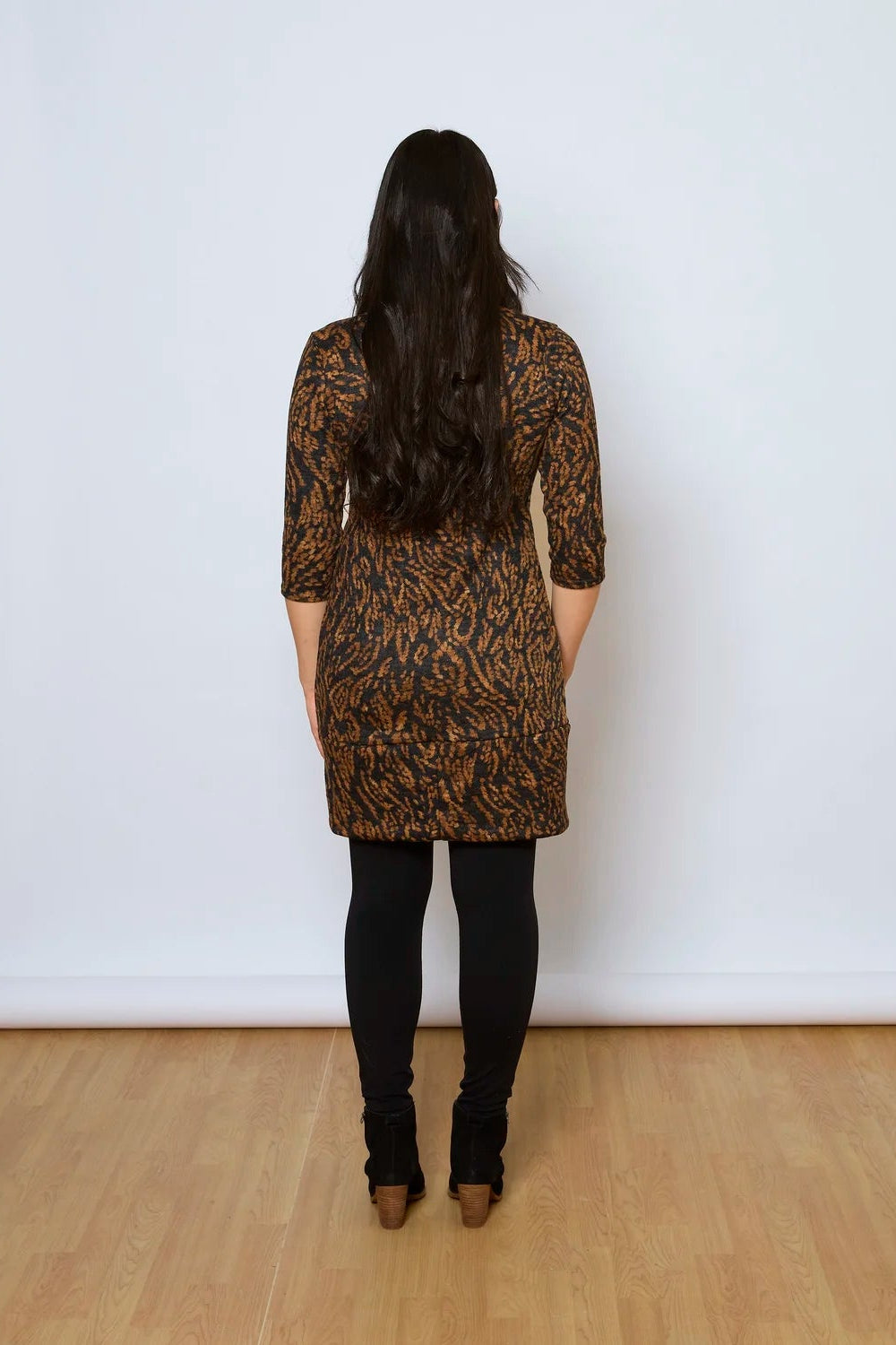 Back view of a woman wearing the Brooke Tunic by Tangente in Rust Print, with 3/4 sleeves and a fitted band at the bottom. She is wearing it with black leggings and standing against a white wall. 