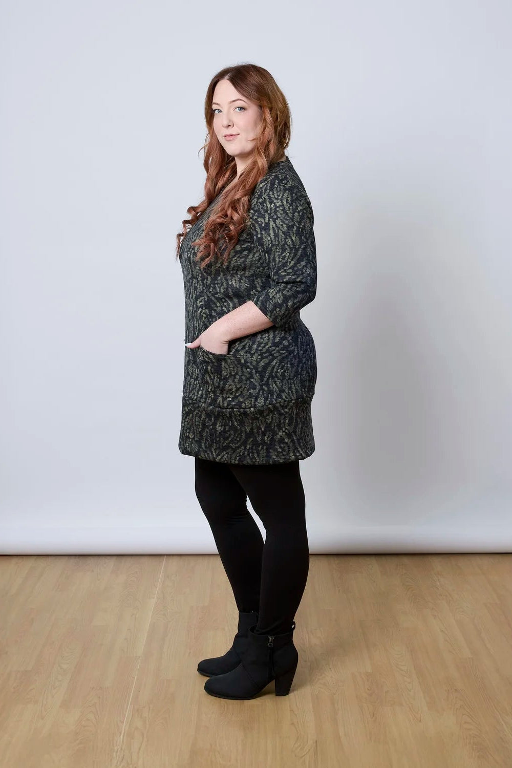 Side view of a woman wearing the Brooke Tunic by Tangente in Green Print, with 3/4 sleeves and a fitted band at the bottom. She is wearing it with black leggings and standing against a white wall. 