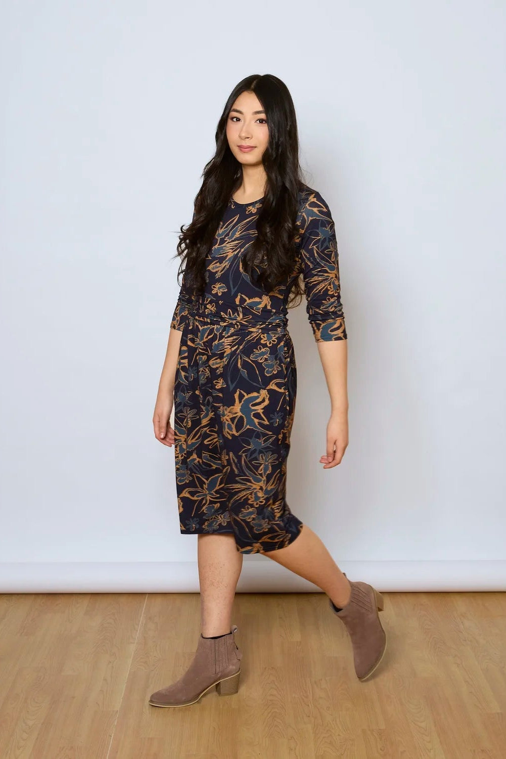 A woman wearing the Cascade Dress by Tangente in Navy Print,  with a round neck, 3/4 sleeves, gathered defining waistband, and a gathered knee-length skirt. She is standing against a white wall.