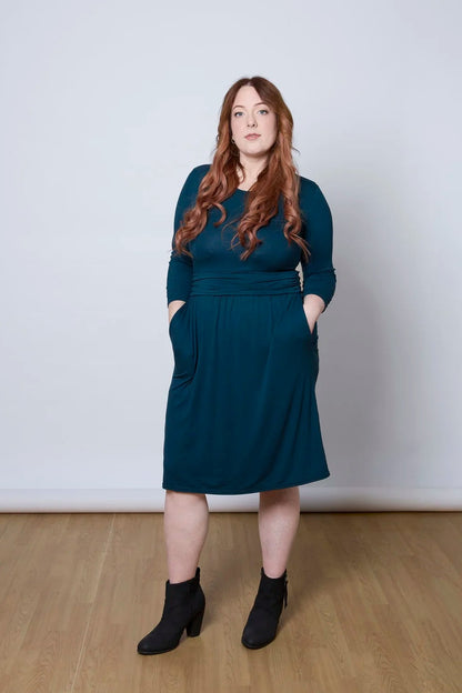 A woman wearing the Cascade Dress by Tangente in Peacock, with a round neck, 3/4 sleeves, gathered defining waistband, and a gathered knee-length skirt. She is standing against a white wall.