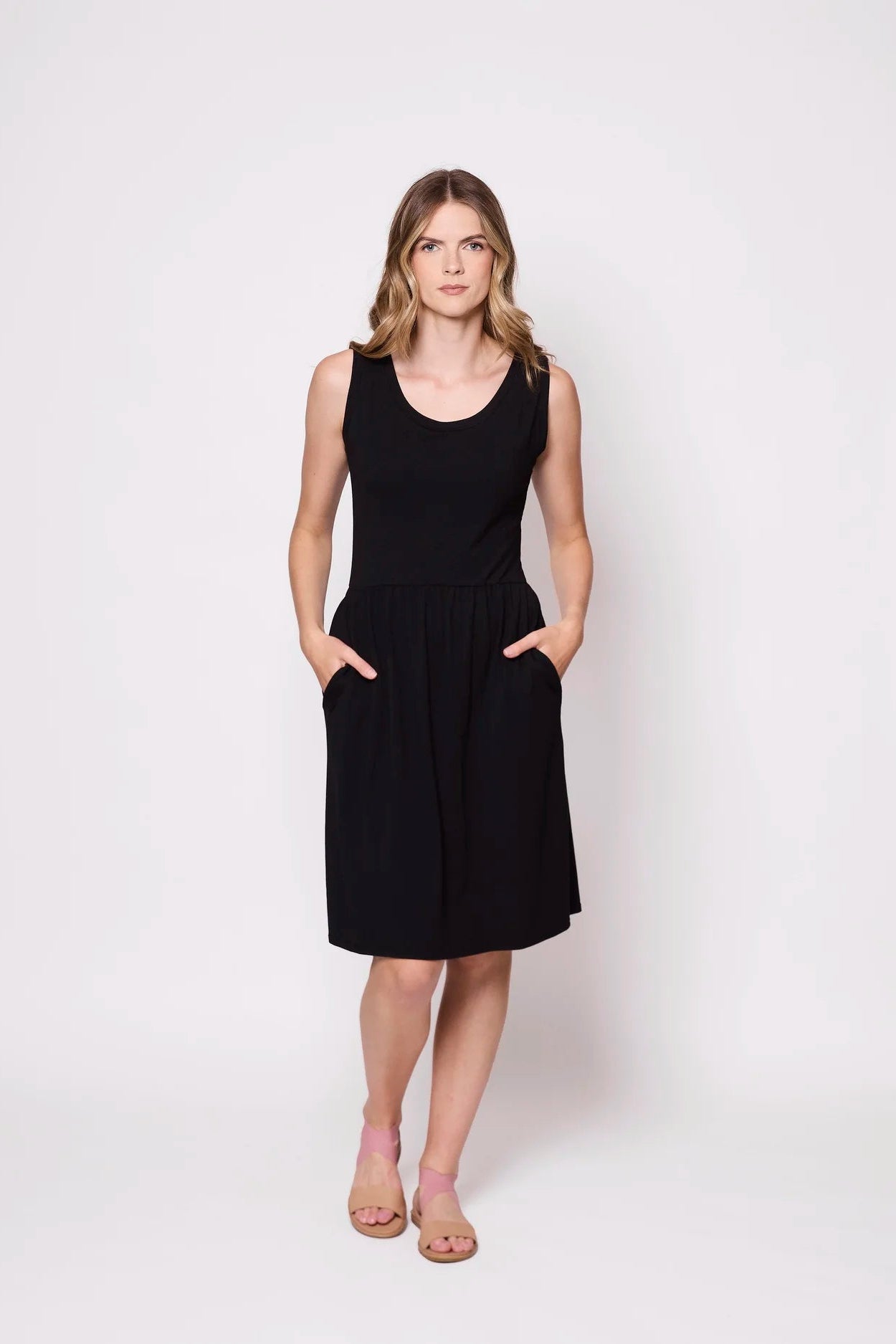 A woman wearing the Mai Dress by Tangente in Black,a  sleeveless dress with a scooped neck and gathered knee-length skirt. She is standing against a white background