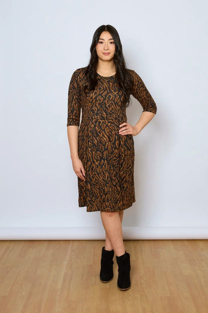 A woman wearing the Meadow Dress by Tangente in Rust Print, with a round neck, 3/4 sleeves, princess seams, and a pleated knee-length skirt. She is standing against a white wall. 