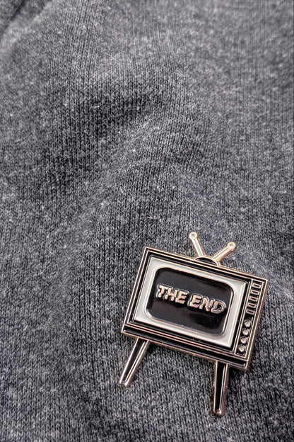  Badaboöm Studio's retro television-shaped enamel pin with 'THE END' lettering on the screen, set against a grey sweatshirt.