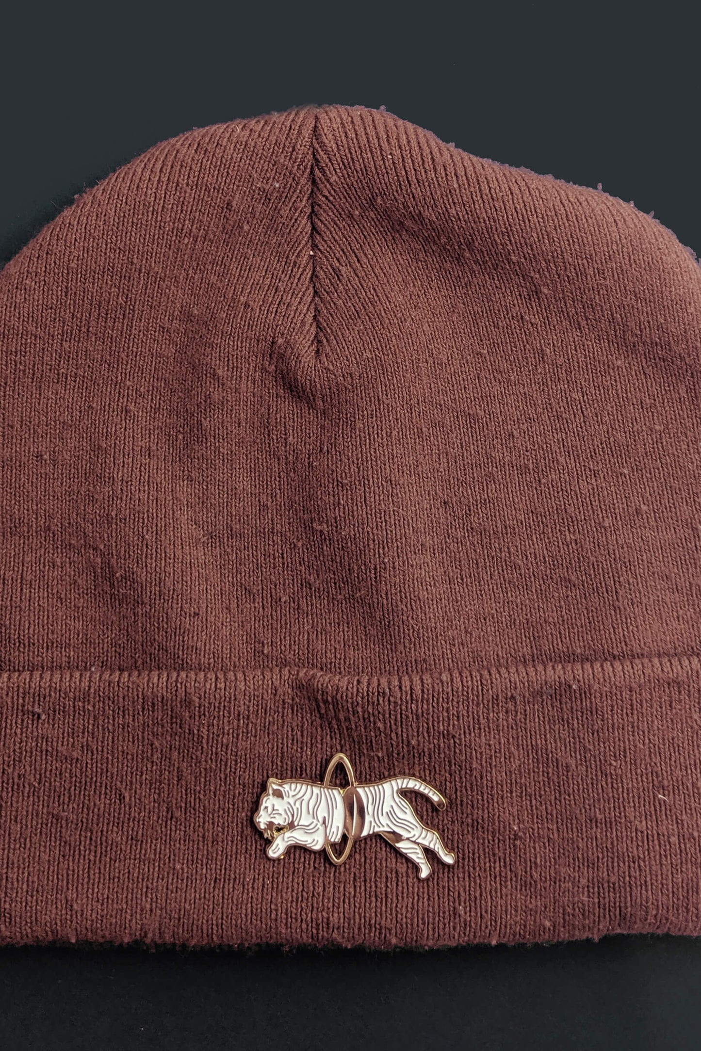 The Tiger Pin from Badaboöm studio, showing a split white and gold tiger leaping through a golden hoop, is shown on a brown toque against a black background. 