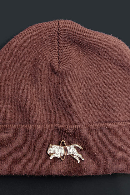 The Tiger Pin from Badaboöm studio, showing a split white and gold tiger leaping through a golden hoop, is shown on a brown toque against a black background. 
