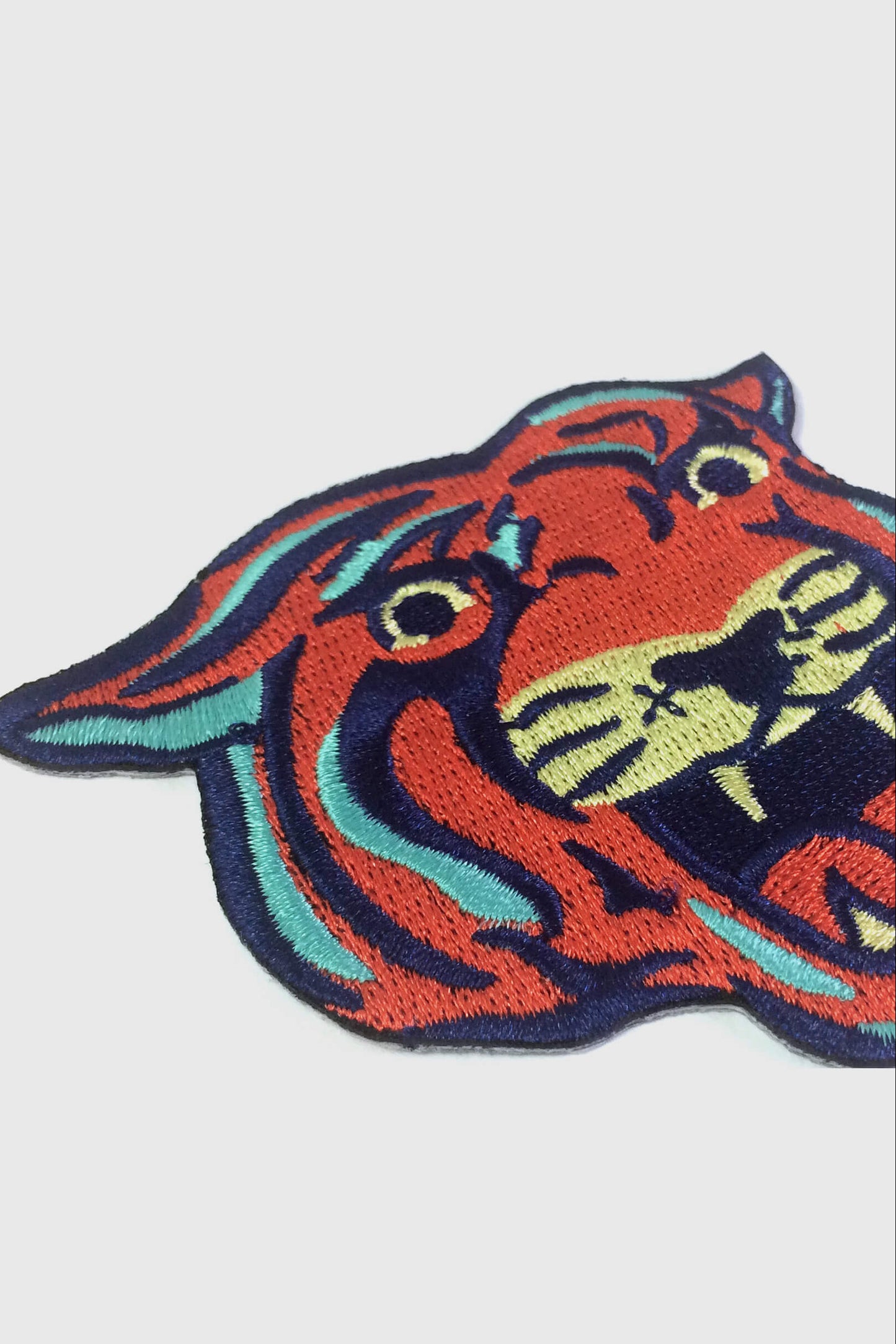 The Tiger embroidered patch from Badaboöm Studio, with orange-red fur, black stripes, and teal and cream accents, centred on a light grey background.
