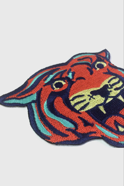 The Tiger embroidered patch from Badaboöm Studio, with orange-red fur, black stripes, and teal and cream accents, centred on a light grey background.