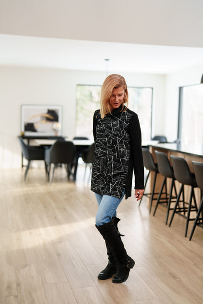 A woman wearing the Auclair Tunic by Rien ne se Perd in a black and white geometric print, with a solid black turtleneck, back, and sleeves, and side slits. She is wearing it with jeans and standing in a room with indoors with a dining room table behind her. 