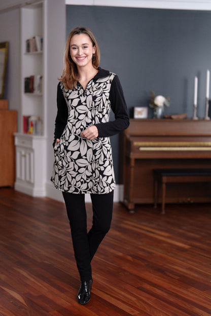 A woman wearing the Lichen Pant by Rien ne se Perd in Black, skinny leg pull-on pants in stretchy corduroy fabric. She is wearing them with a black and white tunic and standing in a room with a piano. 