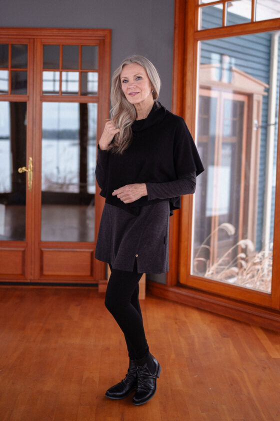 A woman wearing the Modigliani Tunic by Rien ne se Perd in Charcoal and a black poncho standing in front of a floor-to-ceiling window.