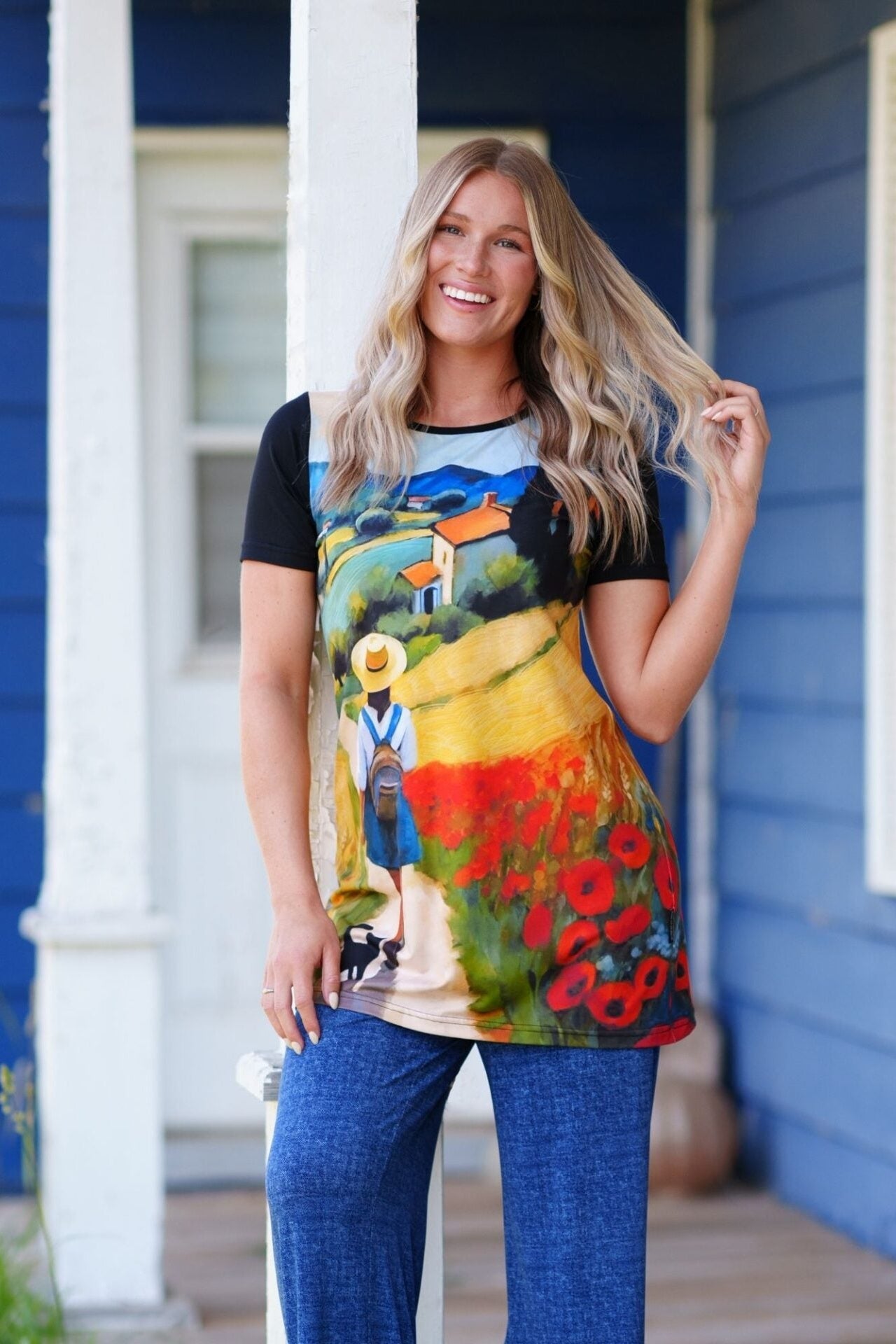 A woman wearing the Negroni Tunic by RNSP, featuring a Tuscan landscape print and contrasting short black sleeves. She is wearing it with jeans and standing outside a blue house. 