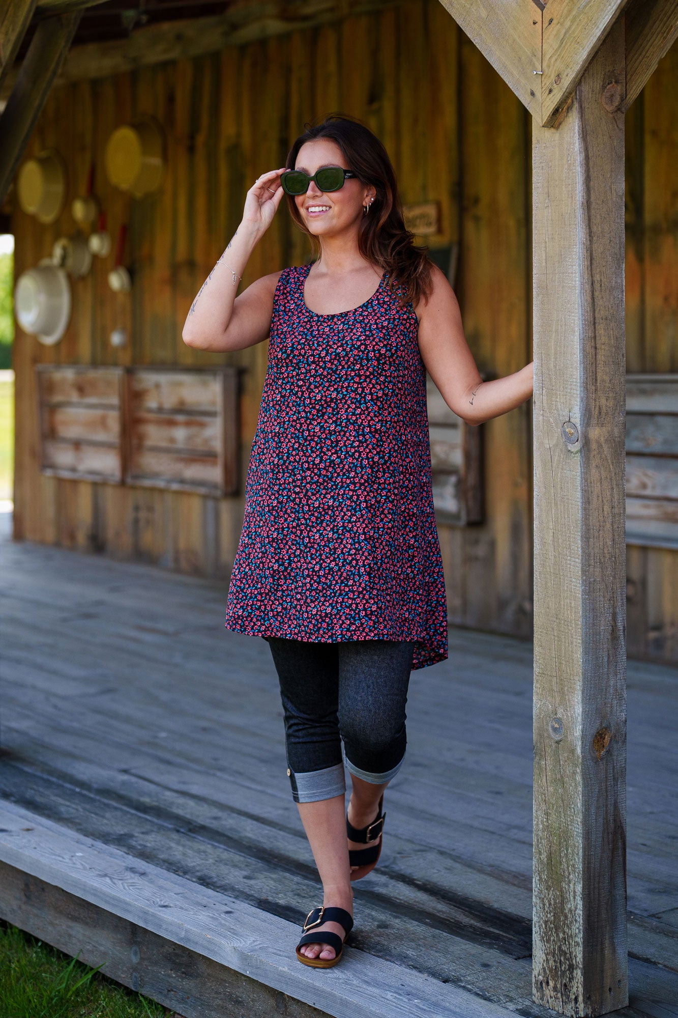 A woman wearing the Oriental Tunic by Rien ne se Perd, featuring a Tiny Flowers print, scoop neck, sleeveless design, trapeze cut, and two pockets. It has a hi-low rounded hemline. She is wearing it with black capri leggings and standing outside in front of a wooden building. 
