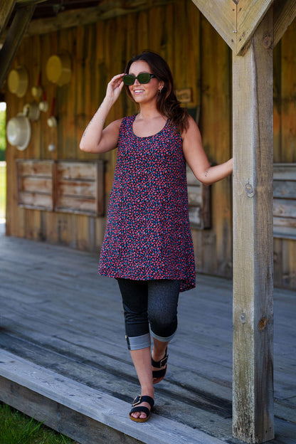 A woman wearing the Oriental Tunic by Rien ne se Perd, featuring a Tiny Flowers print, scoop neck, sleeveless design, trapeze cut, and two pockets. It has a hi-low rounded hemline. She is wearing it with black capri leggings and standing outside in front of a wooden building. 
