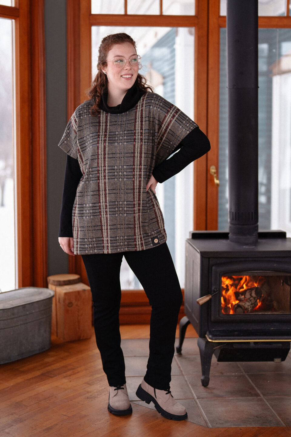 A woman wearing the Lichen Pant by Rien ne se Perd in Black, skinny leg pull-on pants in stretchy corduroy fabric. She is wearing them with plaid top and standing by a fireplace. 