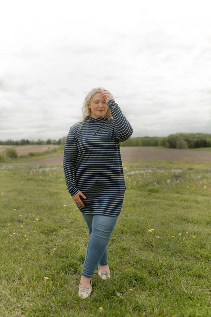A woman wearing the Rupert Tunic by Rien ne se Perd in Navy/Ivory stripe, with  a crossover neckline and hood, long sleeves, and rounded hem. She is wearing it with jeans and standing outdoors in a field.
