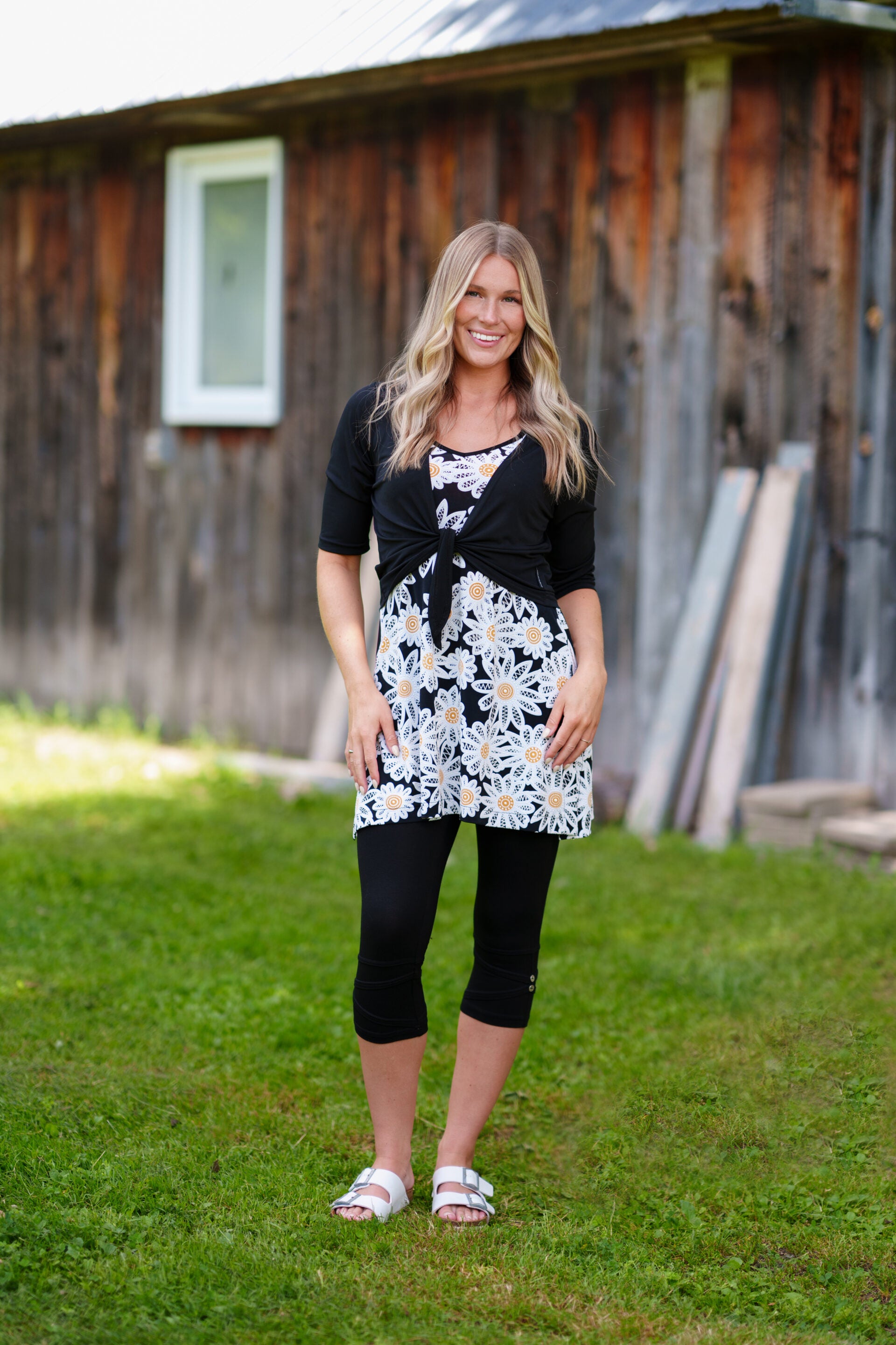 A woman wearing the Petillant Bolero by Rien ne se Perd in Black, with 3/4 sleeves and a tie front. She is wearing it over the Wisky tunic and black leggings  and standing outdoors in front of a wooden building.
