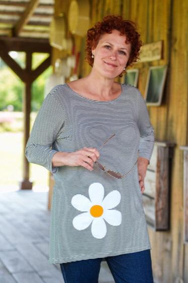 A woman wearing the Zumba Tunic by Rien ne se Perd, a striped navy and white piece with a boat neck, 3/4 sleeves, and a large daisy applique. She is wearing it with jeans and standing outside in from of a wooden building. 