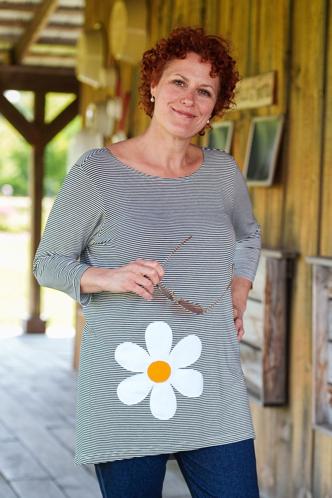 A 3/4 view of a woman wearing the Zumba Tunic by Rien ne se Perd, a striped navy and white piece with a boat neck, 3/4 sleeves, and a large daisy applique. She is wearing it with the Lindy Capri pants and standing outside a wooden building. 