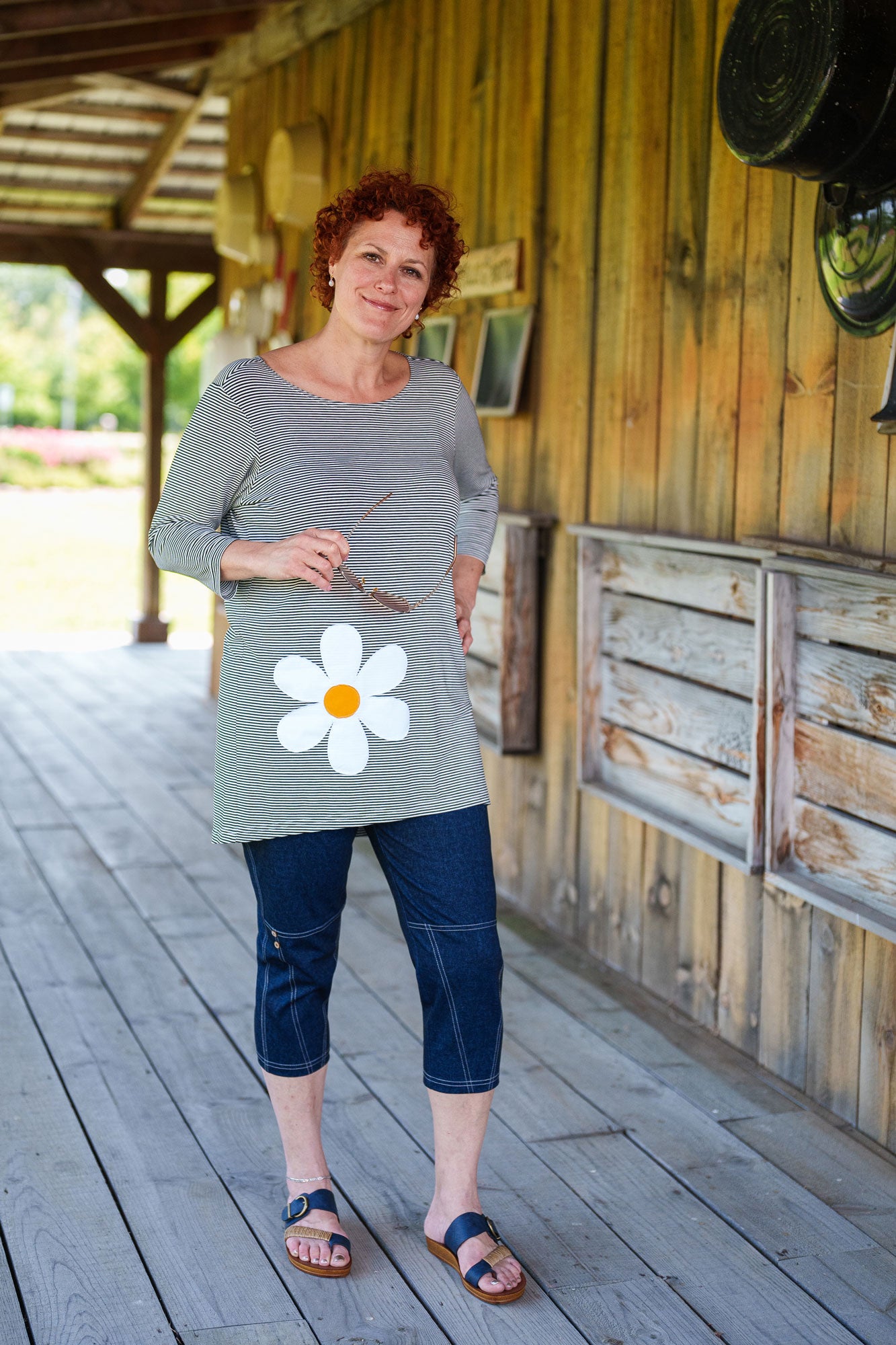 A woman wearing the Zumba Tunic by Rien ne se Perd, a striped navy and white piece with a boat neck, 3/4 sleeves, and a large daisy applique. She is wearing it with the LIndy Capri pants and standing outside a wooden building. 