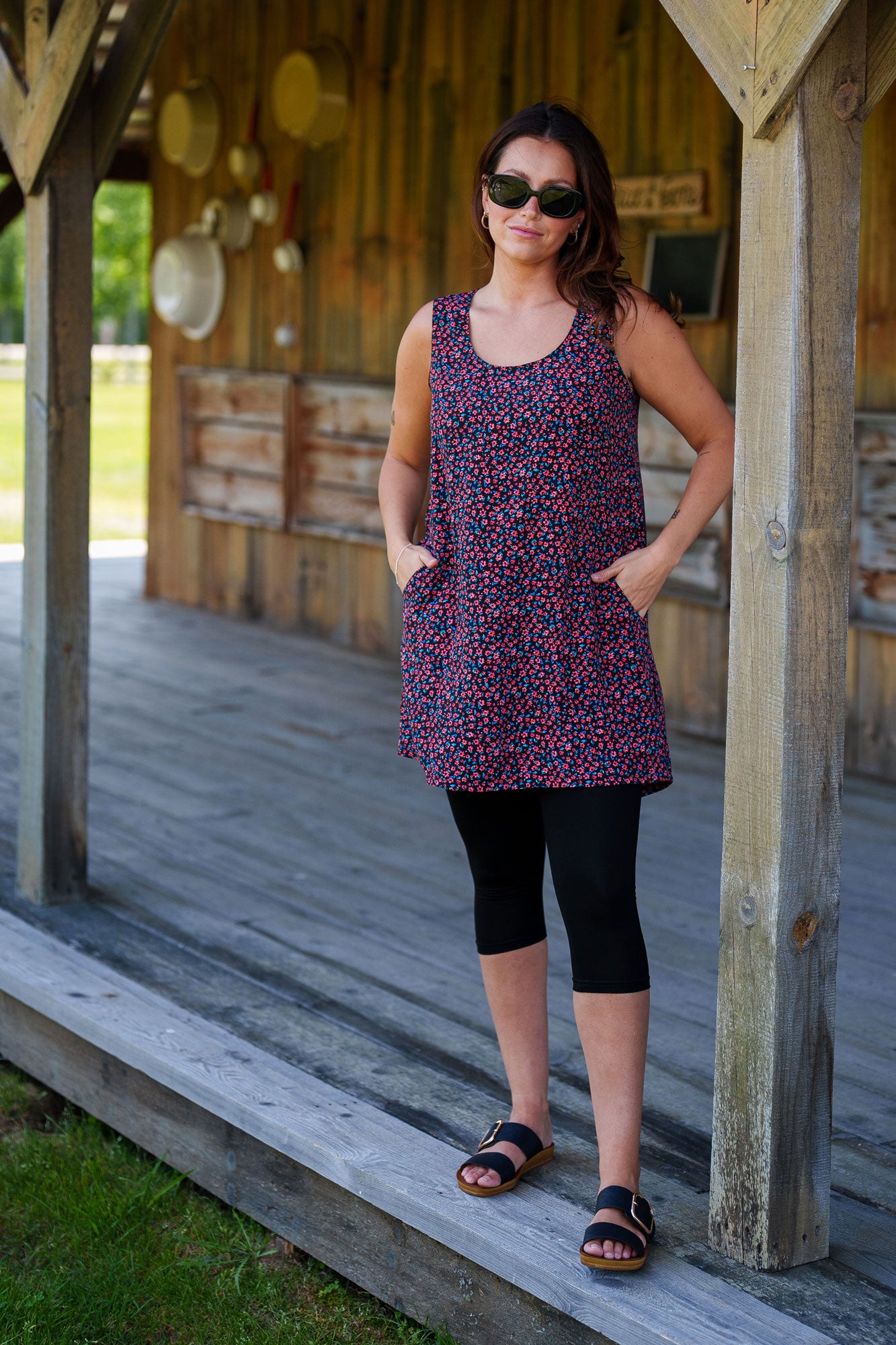 A woman wearing the Oriental Tunic by Rien ne se Perd, featuring a Tiny Flowers print, scoop neck, sleeveless design, trapeze cut, and two pockets. It has a hi-low rounded hemline. She is wearing it with black capri leggings and standing outside in front of a wooden building. 
