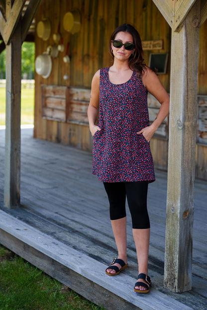 A woman wearing the Oriental Tunic by Rien ne se Perd, featuring a Tiny Flowers print, scoop neck, sleeveless design, trapeze cut, and two pockets. It has a hi-low rounded hemline. She is wearing it with black capri leggings and standing outside in front of a wooden building. 
