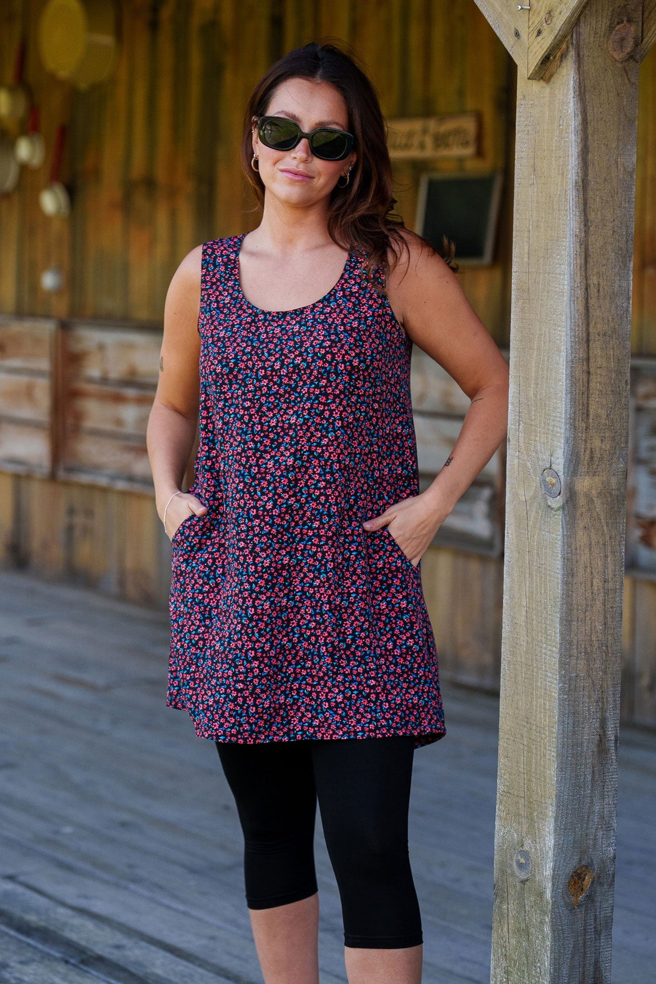 3/4 view ofa woman wearing the Oriental Tunic by Rien ne se Perd, featuring a Tiny Flowers print, scoop neck, sleeveless design, trapeze cut, and two pockets. It has a hi-low rounded hemline. She is wearing it with black capri leggings and standing outside in front of a wooden building. 
