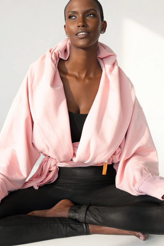 A woman wearing the Heaven Bamboo/Cotton Cropped Wrap from Stacey Martin Lifestyle in Blush Pink, a cropped hoody with a fringed collar scarf that can be tied various ways. She is wearing it with black leggings and seated in a lotus position in a white room. 

