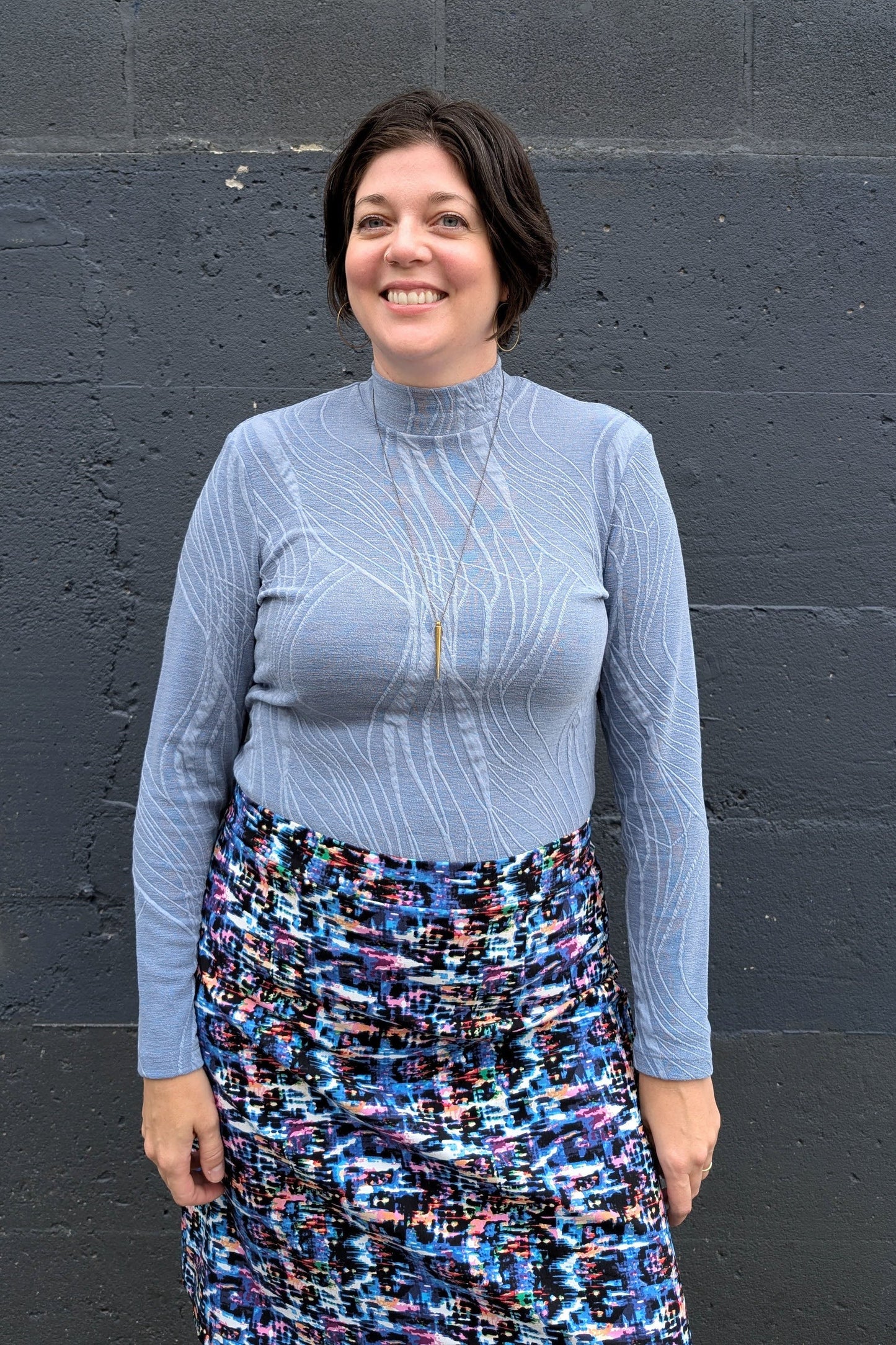 A woman wearing the Franco Mock Top by Luc Fontaine in blue, featuring a textured fabric, mock turtleneck and long sleeves, with the Eve Skirt by Slak, standing outside in front of a brick wall. 