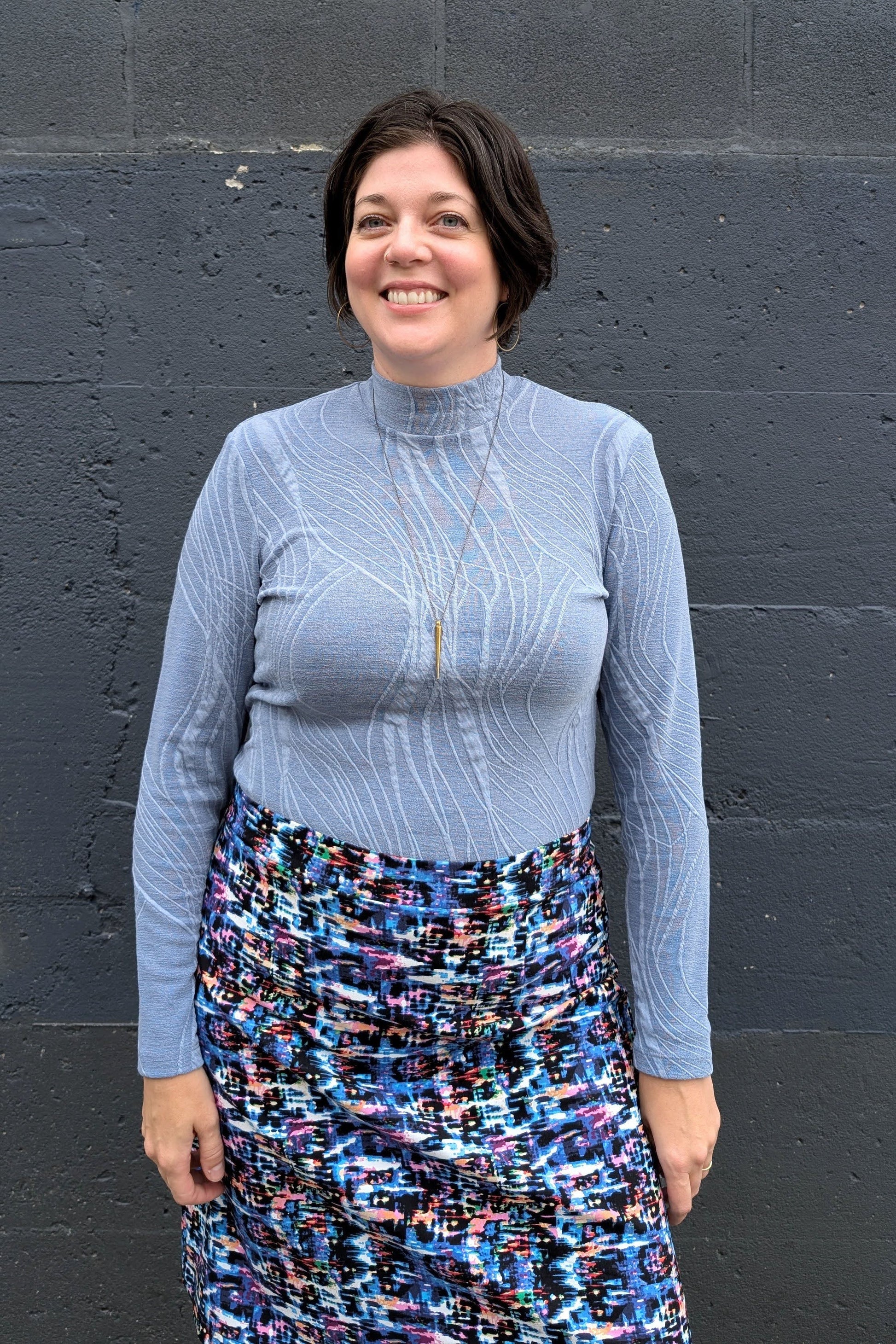 A woman wearing the Franco Mock Top by Luc Fontaine in blue, featuring a textured fabric, mock turtleneck and long sleeves, with the Eve Skirt by Slak, standing outside in front of a brick wall. 