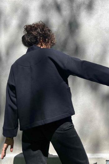 Back view of a woman wearing the Thuya Jacket by Kazak in Black, standing in front of a grey wall 