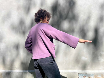 Side view of a woman wearing the Thuya Jacket by Kazak in Purple Haze, standing in front of a grey wall 