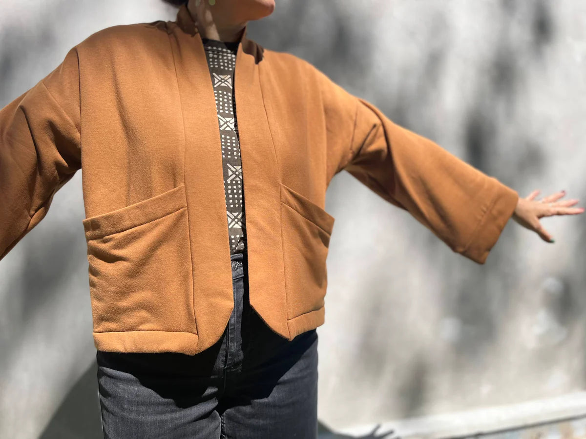 A woman wearing the Thuya Jacket by Kazak in Teak, standing in front of a grey wall 