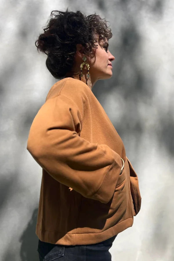 Side view of a woman wearing the Thuya Jacket by Kazak in Teak, standing in front of a grey wall 