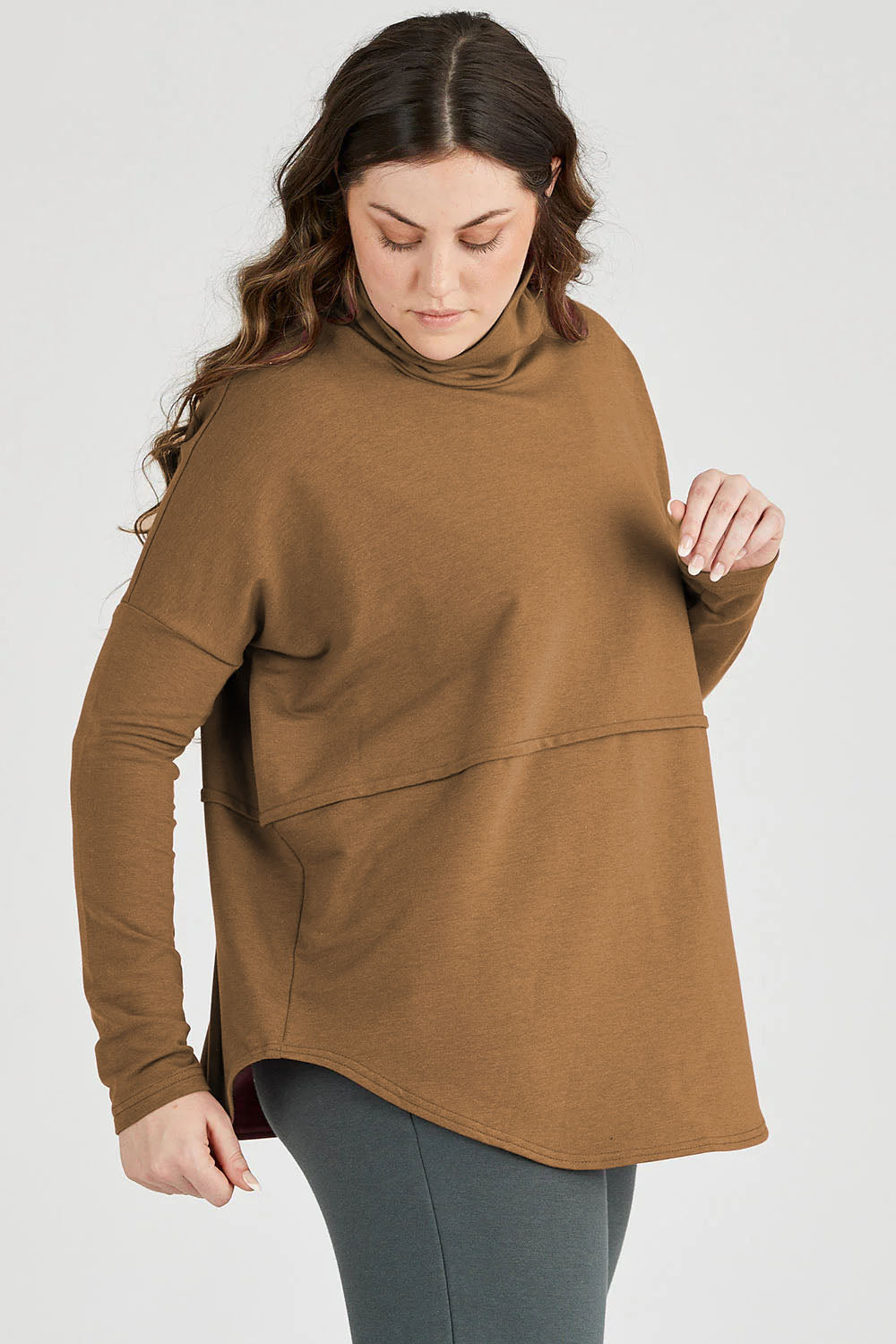 A woman wearing the Abigail Tunic by Advika in Whiskey, featuring a turtleneck, long extended sleeves, a seam across the body, and a rounded hem, in cotton/tencel fabric. She is wearing it with leggings and standing in front of a white background. 