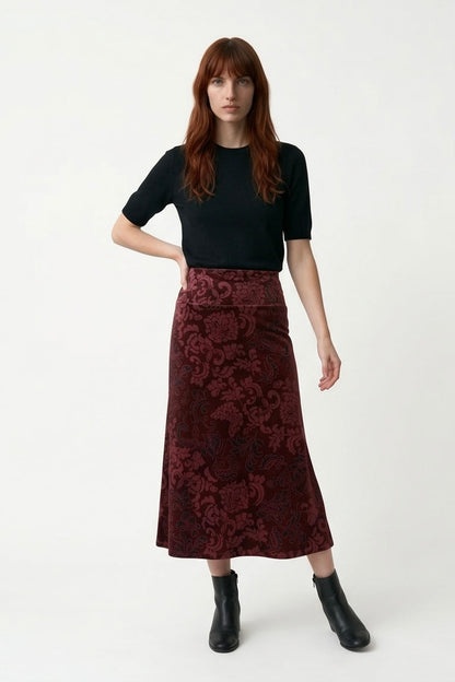 A woman wearing the Kollontai Winslet skirt, a burgundy velvet midi skirt, with a black top. She is standing against a plain white studio background.